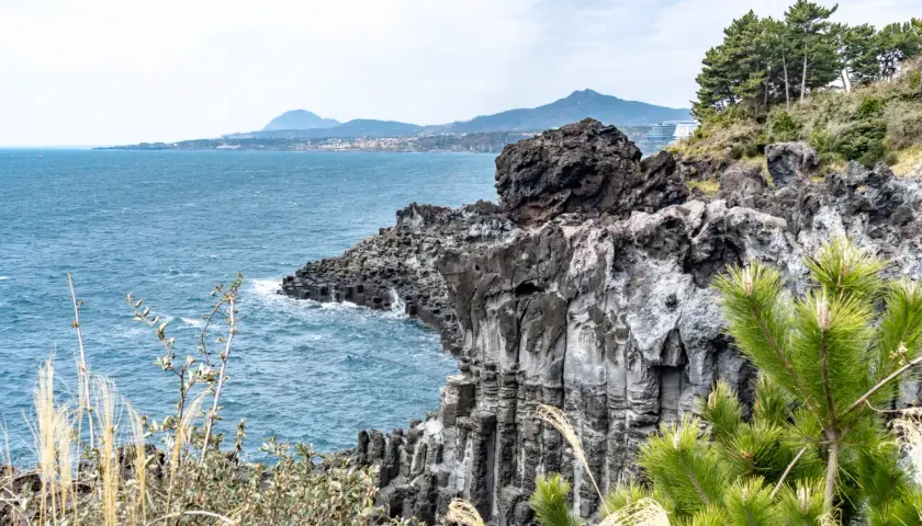 Felsformationen an der Küste der Jeju Insel, mit dem blauen Meer und grünen Bäumen.