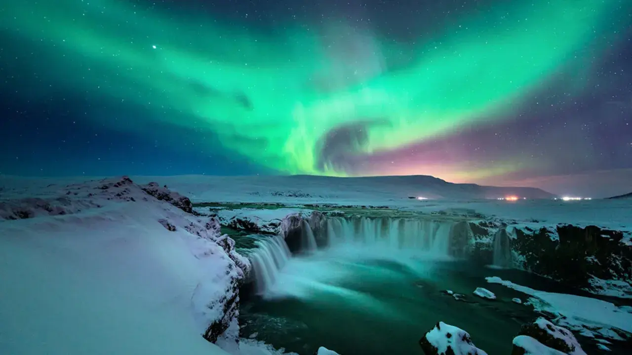 Island reisezeit: Polarlichter tanzen über einem schneebedeckten Wasserfall. Ein magischer Anblick!