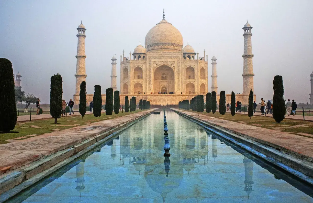 Das Taj Mahal, eine Moschee, spiegelt sich im Wasser. Besucher spazieren durch den Garten.