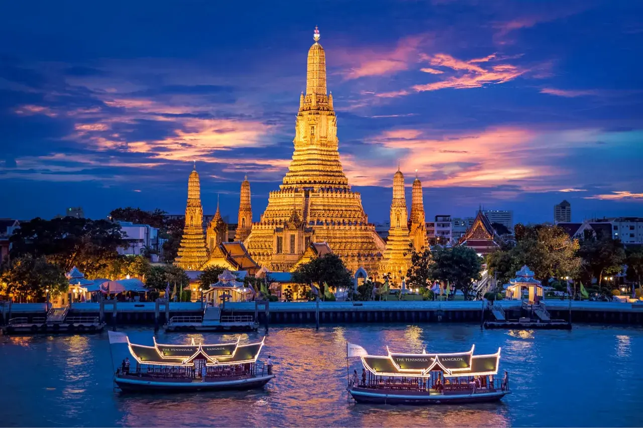 Wat Arun im Abendlicht, ein absolutes Bangkok Must-See. Boote auf dem Fluss vor dem beleuchteten Tempel.