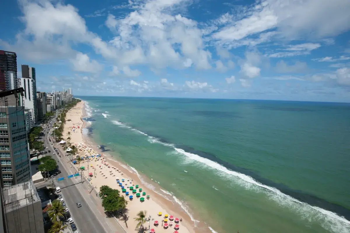 Blick auf den Strand von Recife, Brasilien, mit Palmen, bunten Sonnenschirmen und dem türkisfarbenen Meer unter einem blauen Himmel mit Wolken.