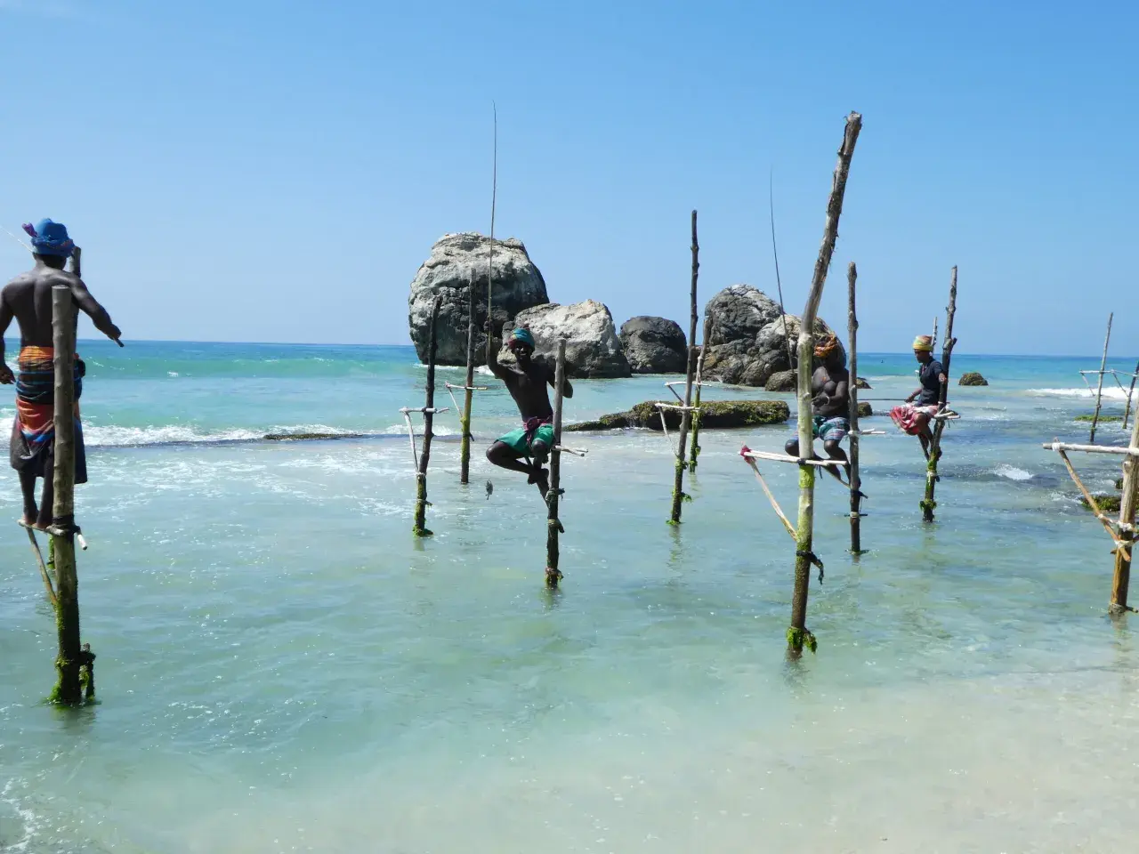 Stelzenfischer Sri Lanka: Männer sitzen auf Stelzen im Meer und angeln. Die Sonne scheint auf das türkisfarbene Wasser.