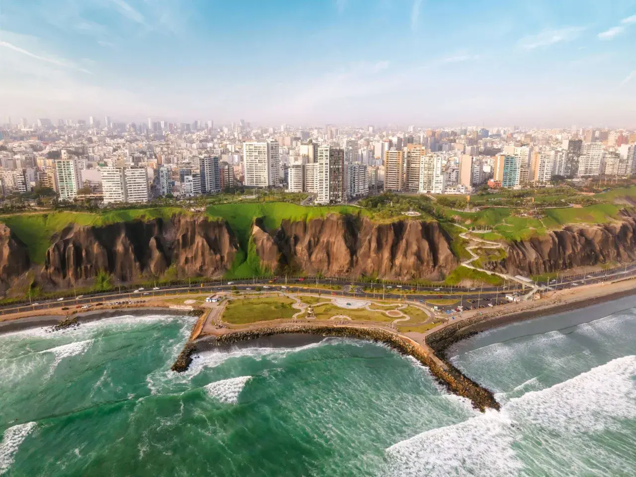 Blick auf die Küste von Lima, wo grüne Hügel auf das Meer treffen. Hochhäuser säumen die Stadt, während die lima einwohner die Küstenpromenade genießen.