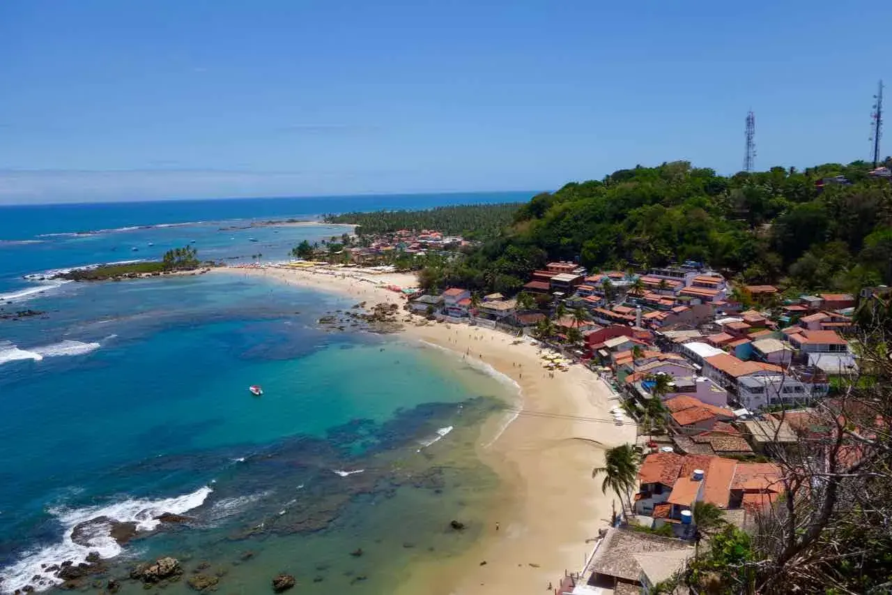 Blick auf die Küste von Morro de São Paulo mit türkisfarbenem Wasser, Sandstrand und bunten Häusern, die sich an einen grünen Hügel schmiegen.
