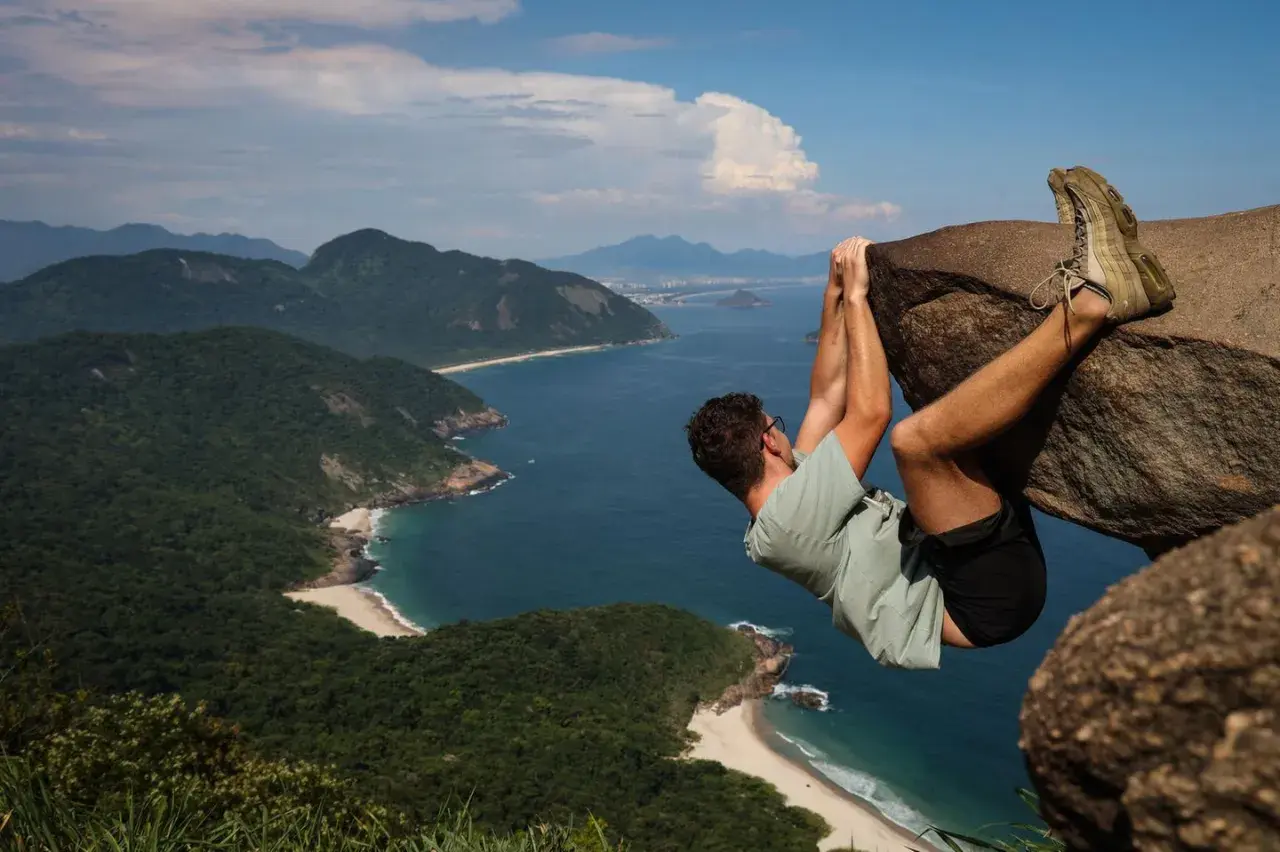 Mann hängt an einem Felsen über dem Meer an der Pedra do Telégrafo. Grüne Hügel und Strände erstrecken sich in der Ferne.