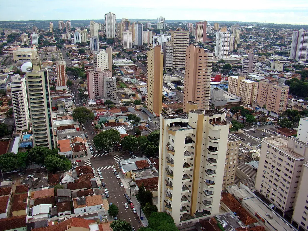 Blick über eine Stadt mit vielen Hochhäusern und kleineren Gebäuden. Ein langer, heller Turm dominiert das Bild, umgeben von weiteren Wohn- und Bürogebäuden. Ein typisches Bild von Campo Grande.