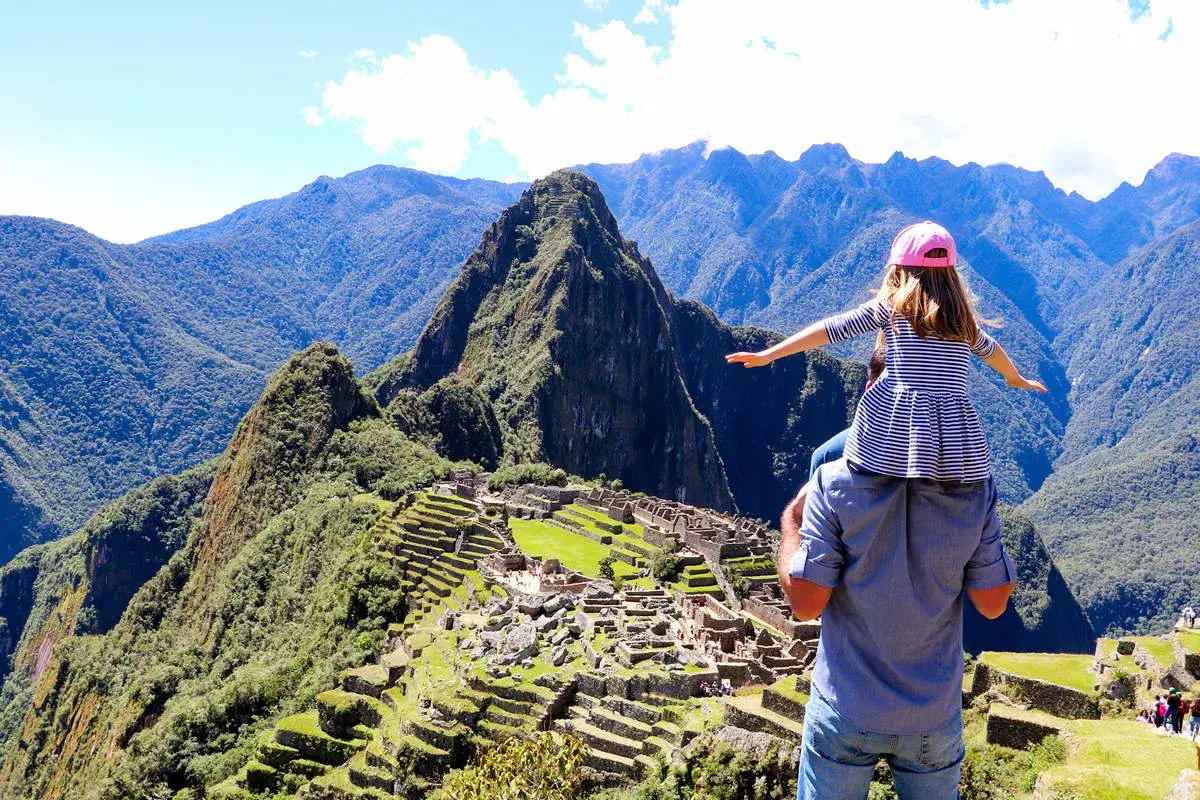 Ein Vater trägt seine Tochter auf den Schultern vor dem majestätischen Machu Picchu Berg. Die antike Stadt liegt eingebettet in die grüne Berglandschaft.