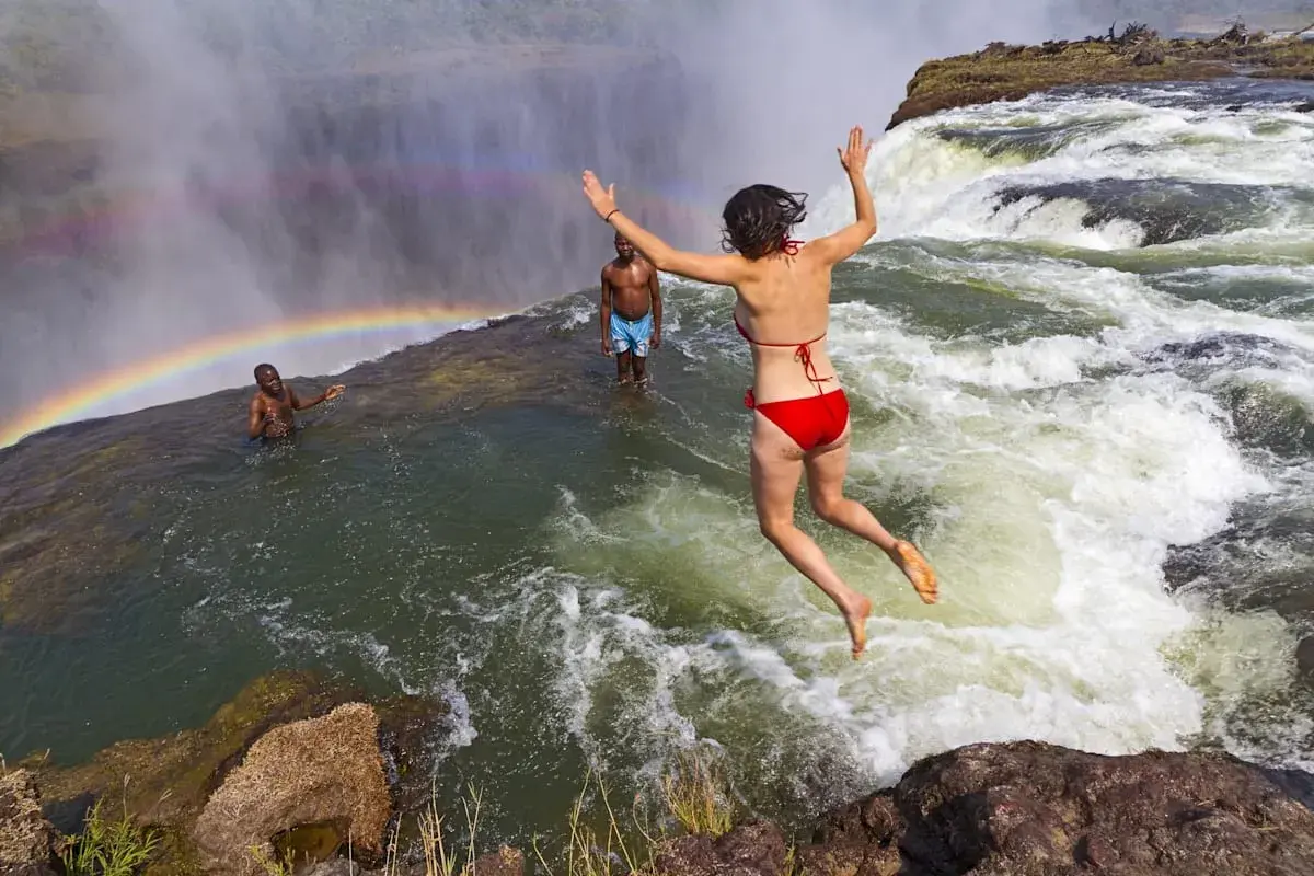Mutige Frau springt in den Devils Pool am Rand der Victoriafälle, ein Regenbogen bildet sich im Sprühnebel.