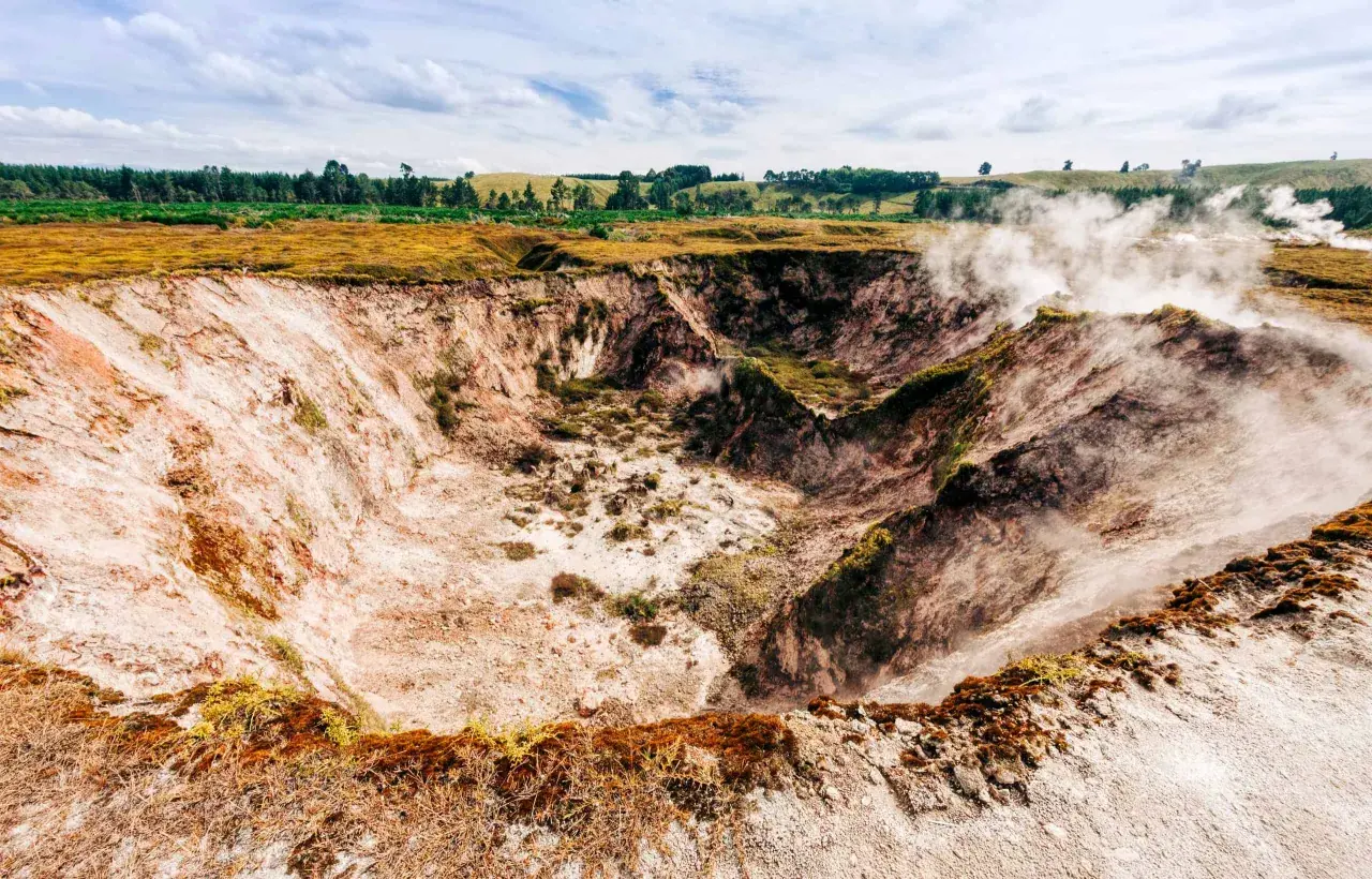 Dampf steigt aus einem Krater in Taupō, Neuseeland. Die Erde ist rötlich-weiß, mit grüner Vegetation am Rand.