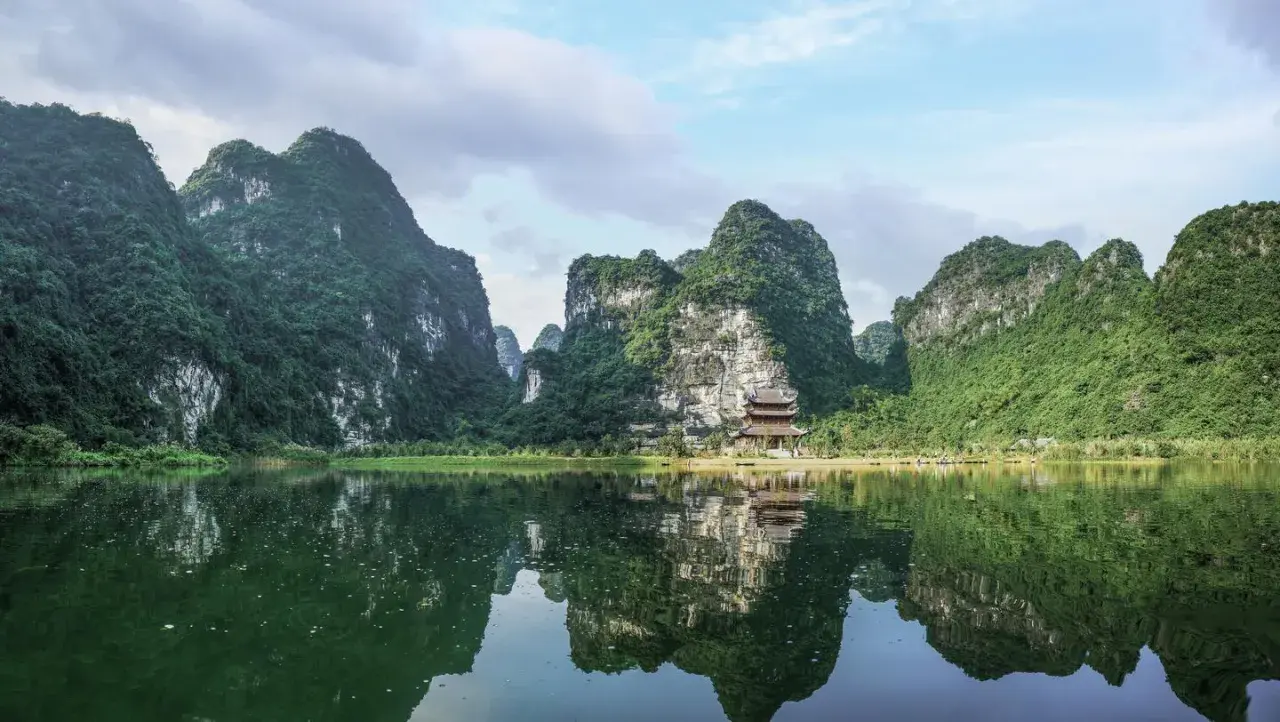 Grüne Berge spiegeln sich im ruhigen Wasser. Ein Tempel schmiegt sich an den Hang. Perfekt für die beste Reisezeit Vietnam.