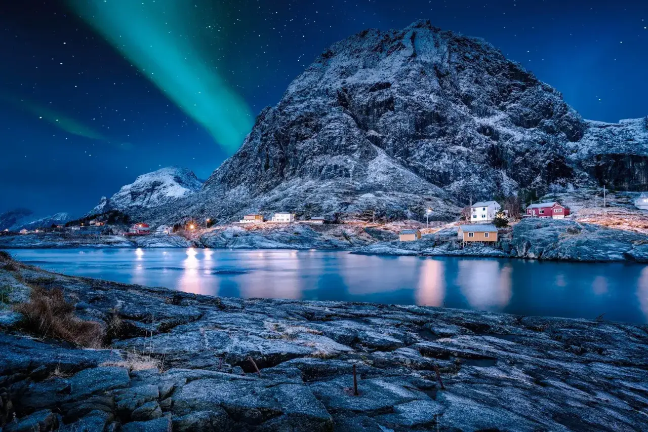 Nordkap Polarlichter tanzen über verschneite Berge und kleine Häuser am Fjord. Ein magischer Anblick!