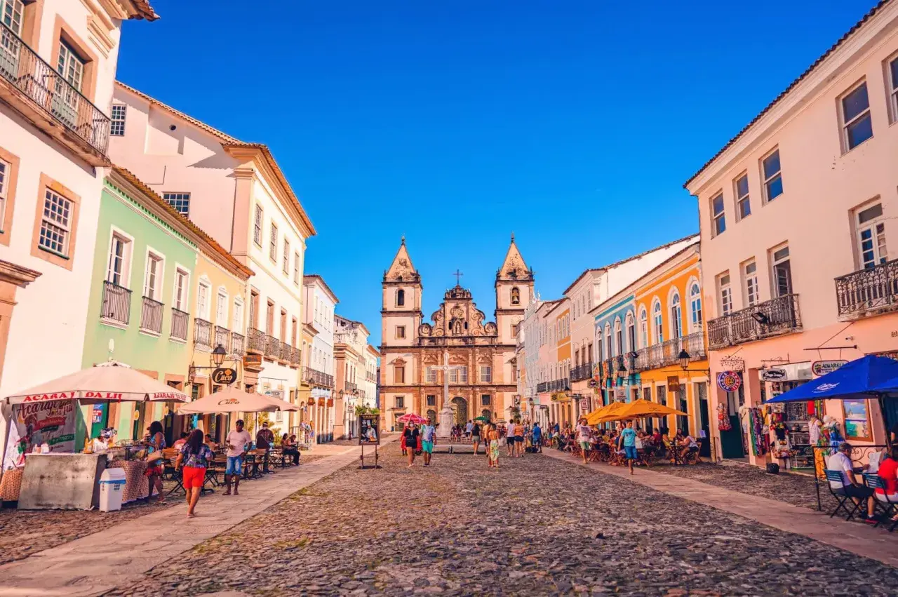 Bunte Kolonialhäuser säumen eine Kopfsteinpflasterstraße in Salvador de Bahia. Menschen flanieren, genießen die Sonne und besuchen Geschäfte.