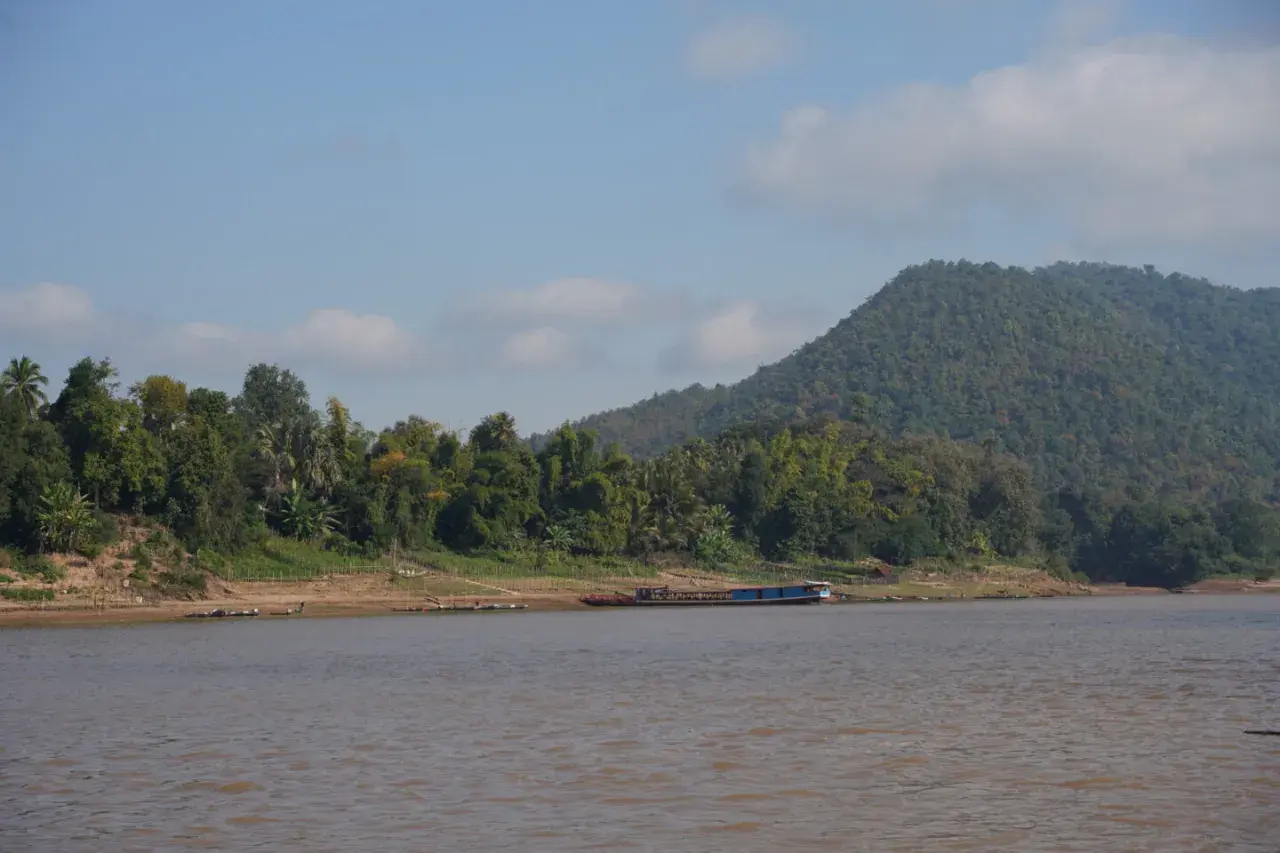 Ein breiter, träger Fluss in Laos, gesäumt von üppiger Vegetation und einem bewaldeten Hügel. Ein langes, blaues Boot liegt am Ufer.