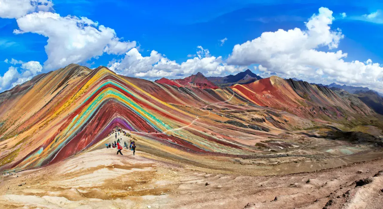 Regenbogenberg in Peru im September. Wanderer bestaunen die bunten Mineralstreifen der Berge unter blauem Himmel.