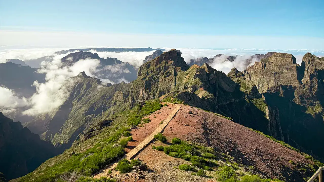 Atemberaubende Berglandschaft auf Madeira, mit Wolken, die sich durch zerkl&uuml;ftete Gipfel schl&auml;ngeln. Ein Pfad f&uuml;hrt &uuml;ber einen roten Hang, ein wahres Highlight von Madeira.