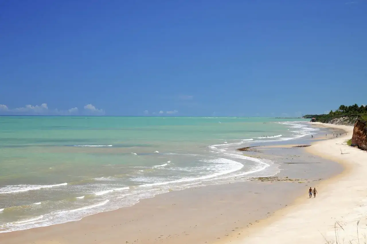 Ein Paar spaziert am Strand von Porto Seguro, Bahia. Türkisfarbenes Meer trifft auf goldenen Sand unter blauem Himmel.