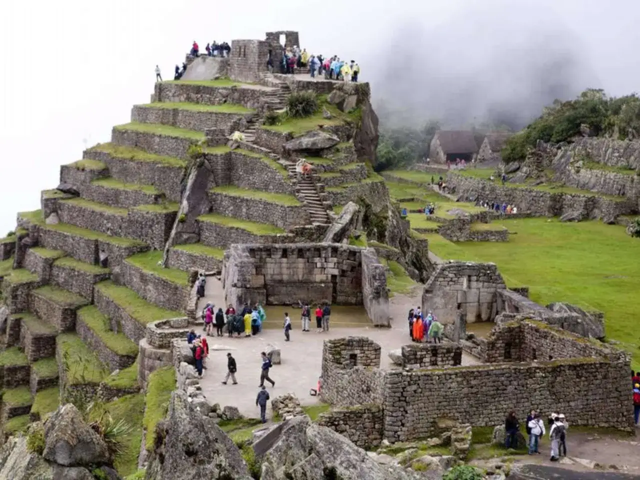 Besucher erkunden die beeindruckende Machu Picchu Rekonstruktion mit ihren terrassierten Feldern und Steingebäuden, umgeben von nebligen Bergen.