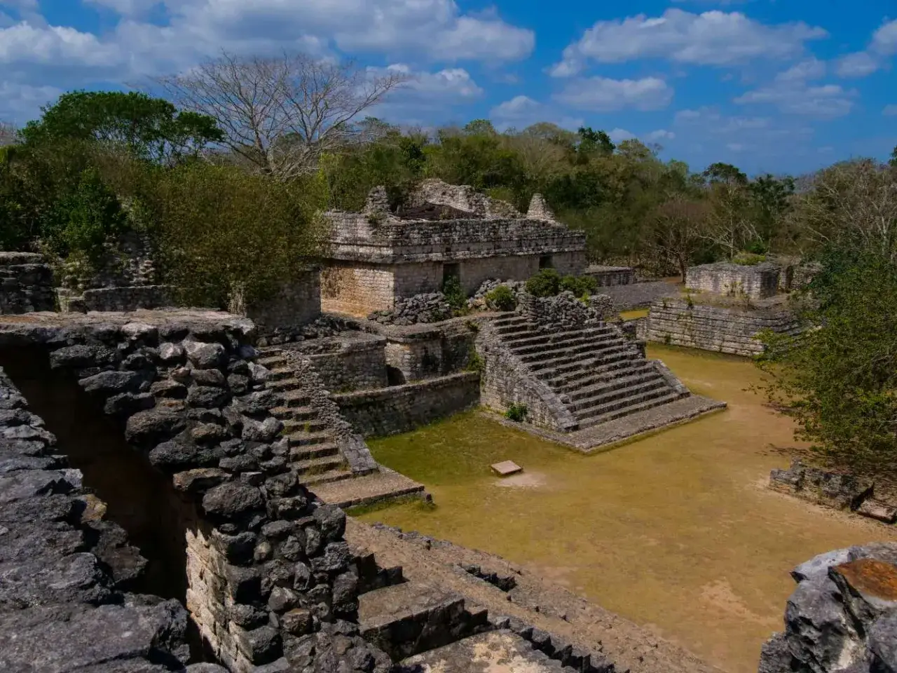 Ruinen einer alten aztekischen Pyramide, umgeben von üppiger Vegetation und unter einem blauen Himmel mit Wolken.