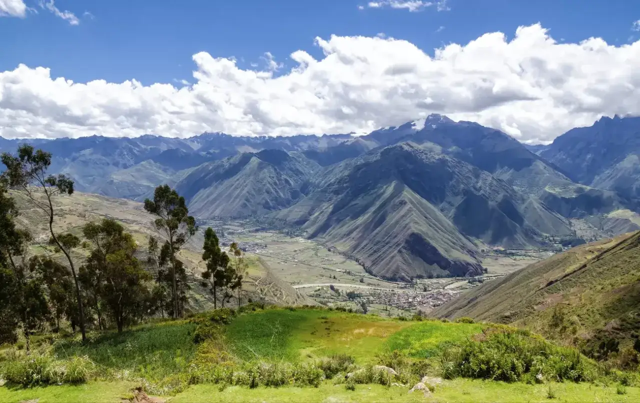 Gr&uuml;ne Terrassenfelder und B&auml;ume blicken auf ein Dorf im Tal der Peru Anden. Majest&auml;tische Berge erheben sich im Hintergrund unter einem blauen Himmel mit wei&szlig;en Wolken.