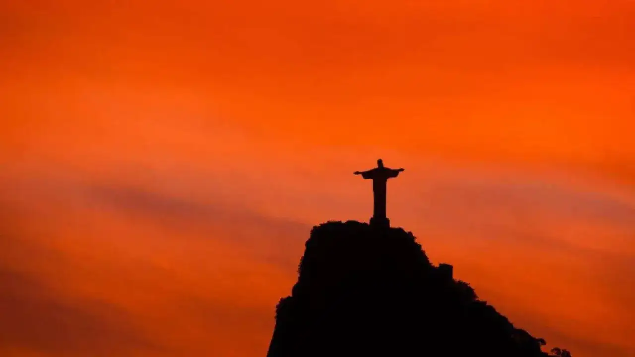 Die ber&uuml;hmte Christusstatue in Rio de Janeiro ragt silhouettenhaft vor einem leuchtend orangen Himmel auf.
