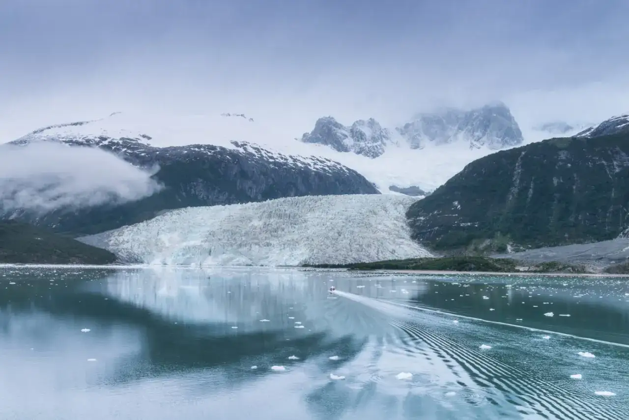 Majestätischer Gletscher in der Patagonia. Ein kleines Boot hinterlässt eine Spur auf dem ruhigen Wasser, umgeben von Eisbergen und schneebedeckten Bergen.