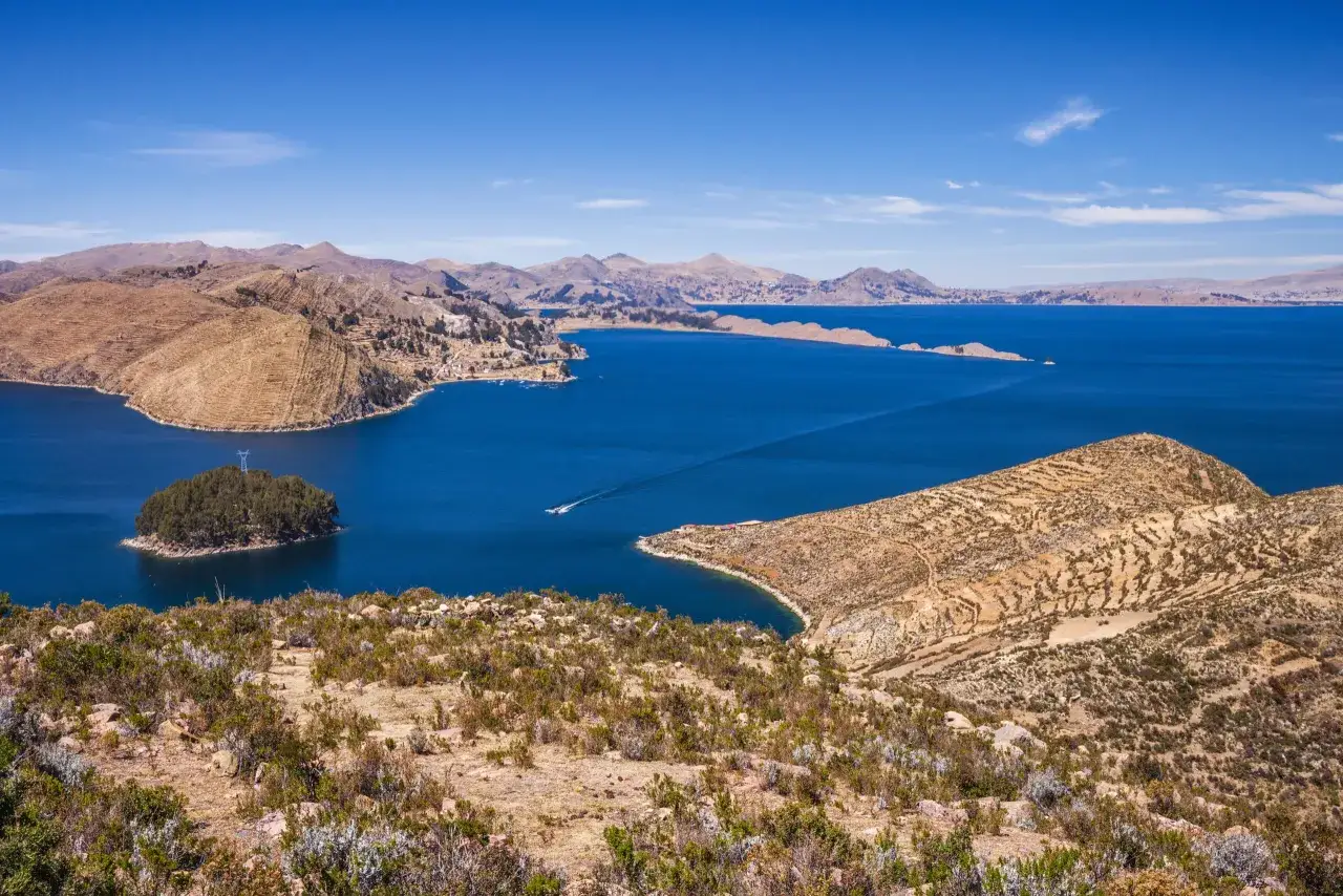Blick auf den Titicacasee, wo er liegt: eine weite Wasserfläche umgeben von trockenen, terrassierten Hügeln und einer kleinen Insel mit Bäumen.
