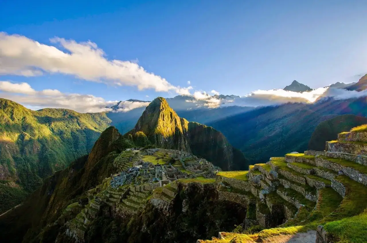 Sonnenaufgang über Machu Picchu, einem der Peru-Highlights. Terrassen und Ruinen schmiegen sich an grüne Berge unter blauem Himmel mit Wolken.