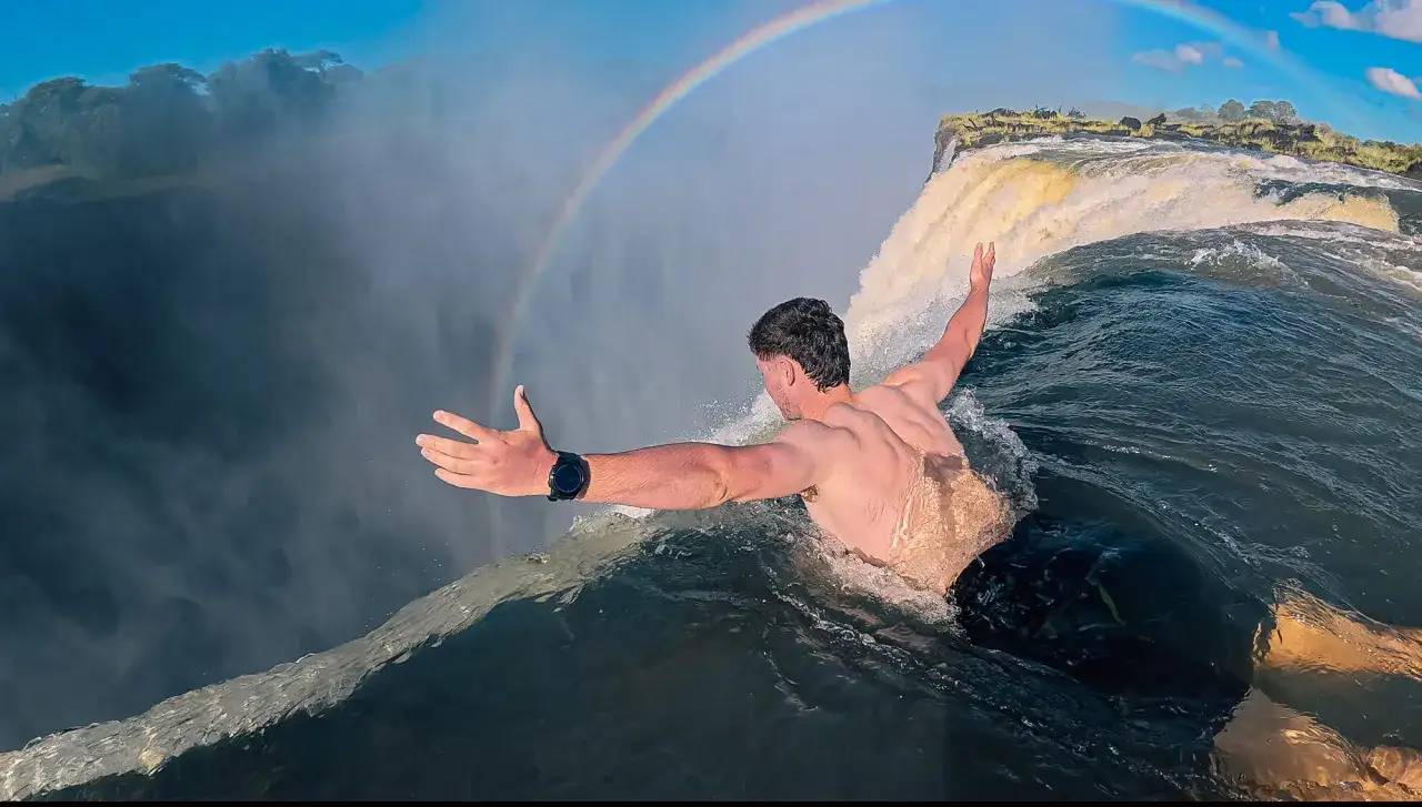 Mann schwimmt im Devils Pool am Rand der Victoriafälle, ein Regenbogen spannt sich über die Gischt.