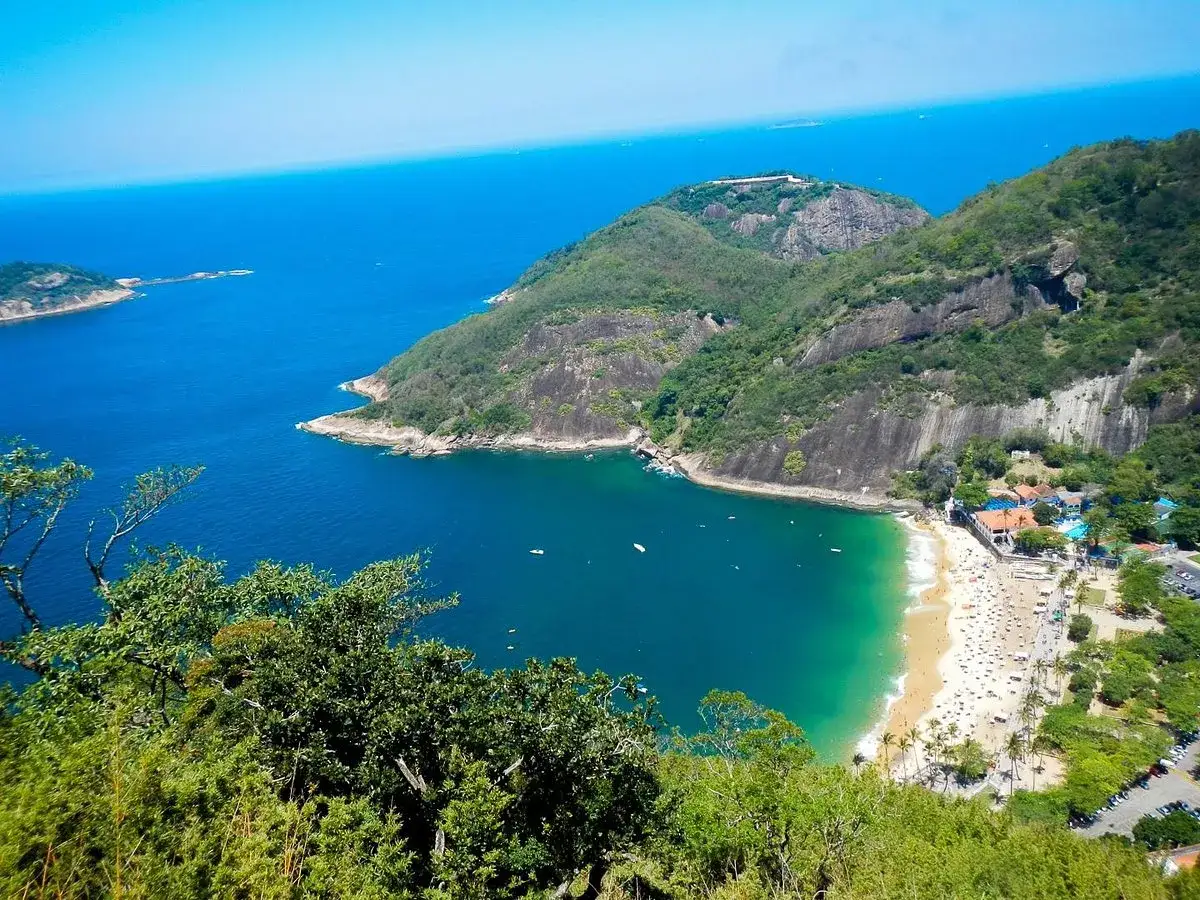 Blick vom Morro da Urca auf die Bucht mit einem belebten Strand, türkisfarbenem Wasser und grünen Hügeln.