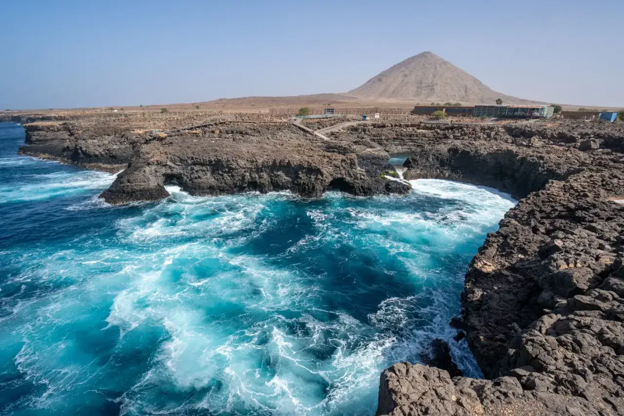 T&uuml;rkisblaues Meer brandet an felsige K&uuml;sten auf den Kapverden im Juli. Im Hintergrund ragt ein vulkanischer Berg auf.