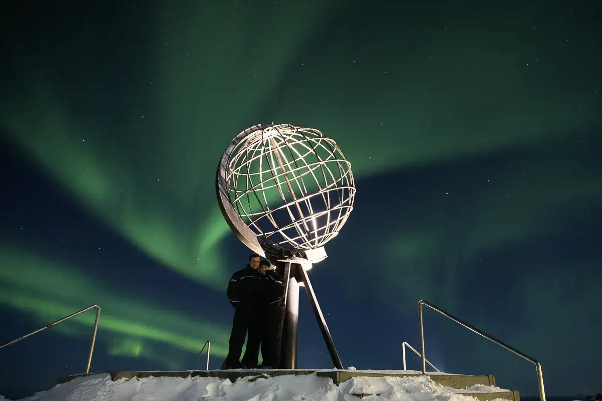 Zwei Personen stehen vor dem Globus am Nordkap, während die Nordlichter den Nachthimmel in Grün tauchen.