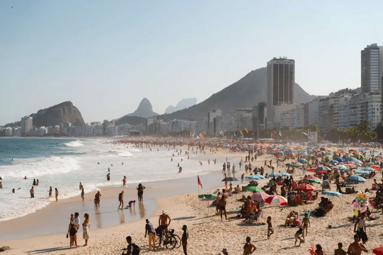 Leblon, Rio de Janeiro: Ein sonniger Tag am Strand mit vielen Menschen, bunten Sonnenschirmen und dem ikonischen Zuckerhut im Hintergrund.