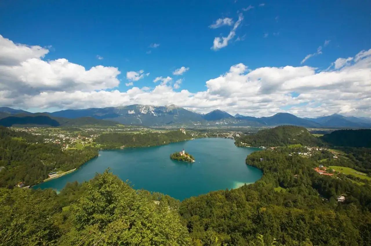 Der Bleder See mit seiner Inselkirche und der Burg, umgeben von grünen Hügeln und majestätischen Bergen, unter einem blauen Himmel mit Wolken.