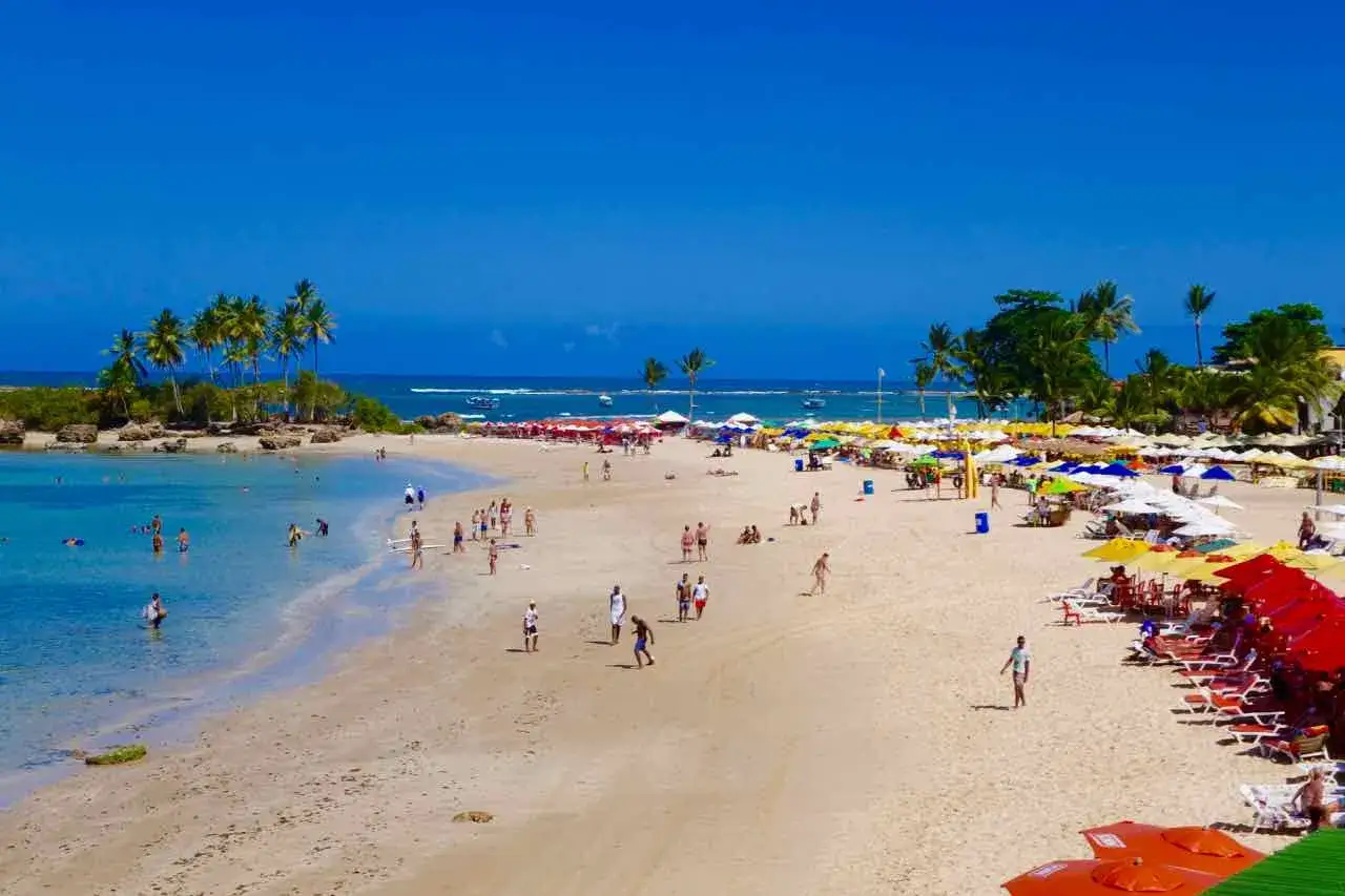 Sonniger Strand von Morro de São Paulo mit vielen bunten Sonnenschirmen, Palmen und Menschen, die im türkisfarbenen Wasser planschen.