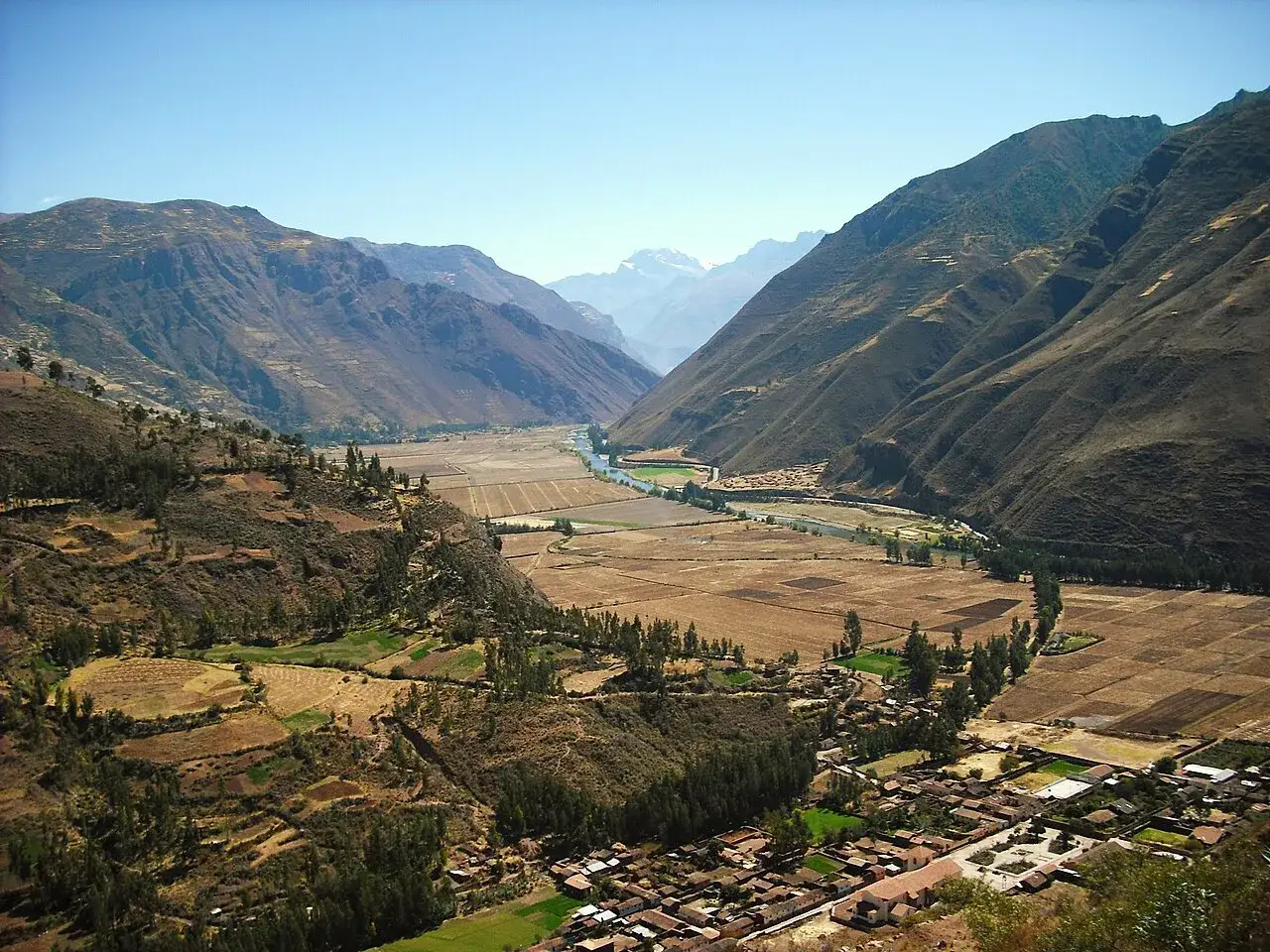 Weites Tal mit Fluss und Feldern im heiligen Tal Peru. Berge und ein Dorf säumen die Landschaft.