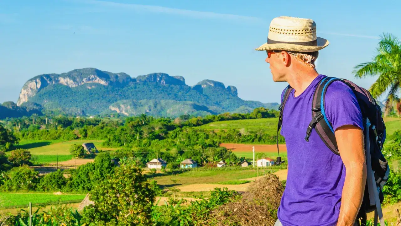 Ein Mann mit Hut und Rucksack blickt auf die grüne Landschaft von Viñales, Kuba. Berge und kleine Häuser im Tal.